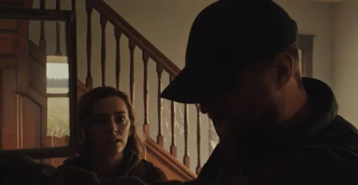 A man stands looking into a cabinet with a class door, a woman stands to his right looking at him under a staircase in THINGS WILL BE DIFFERENT