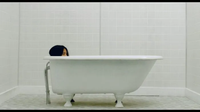 A young woman's head peeks out from a bathtub in an tirely white tiled room