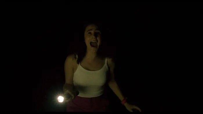 A young woman holding a flashlight is seen screaming in the dark