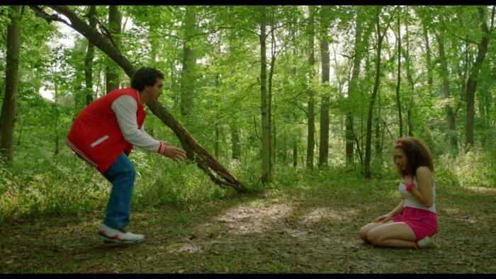 A man in a letterman jacket backs away into the woods while speaking to a woman on the leaf-layered ground.