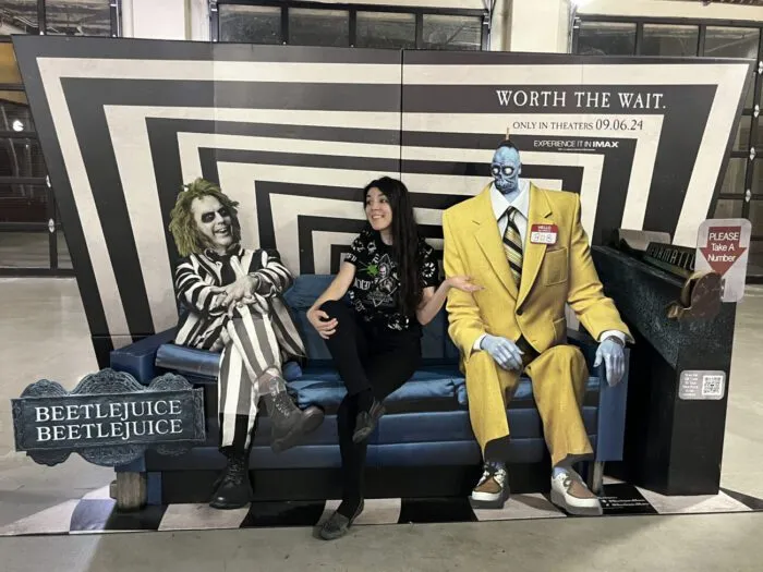 Jamie Lee, in a “Beetlejuice Beetlejuice” T-shirt with a “Beetlejuice” button-up shirt over it, sits on a couch in a waiting room talking to Beetlejuice (Michael Keaton) while sitting between him Bob in a promo standee for the film, “Beetlejuice Beetlejuice” (2024).