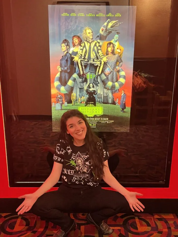 Jamie Lee, in a “Beetlejuice Beetlejuice” T-shirt with a “Beetlejuice” button-up shirt over it, smiles below a poster for “Beetlejuice Beetlejuice” (2024), in the same pose as Beetlejuice (Michael Keaton) on the poster.