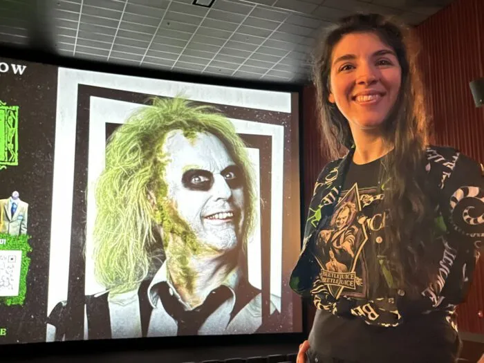 Jamie Lee, in a “Beetlejuice Beetlejuice” T-shirt with a “Beetlejuice” button-up shirt over it, smiles in front of a screen showing a promo image of Beetlejuice (Michael Keaton) before an early screening of “Beetlejuice Beetlejuice” (2024).