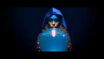 A blue hooded woman with glittery makeup over her forehead and eyes, extends her arms toward the camera holding out a fishbowl filled with water in SCARLET BLUE
