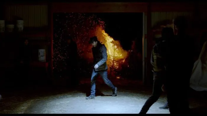 A man walks through a barn where an open door spies a fire raging outside.