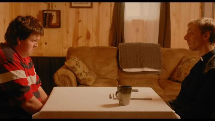 A young man sits opposite a priest at a table in a cabin.