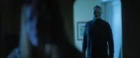 A masked man stands in a lit doorway behind an out of focus woman in He Never Left