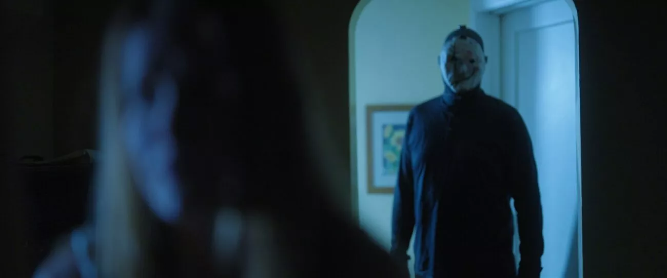 A masked man stands in a lit doorway behind an out of focus woman in He Never Left