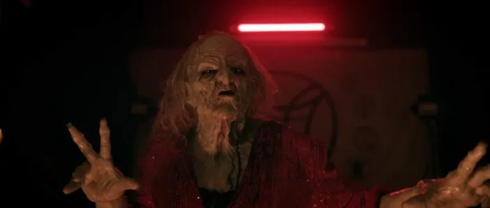 A witch with a wrinkly face stands under a neon light in Members Club