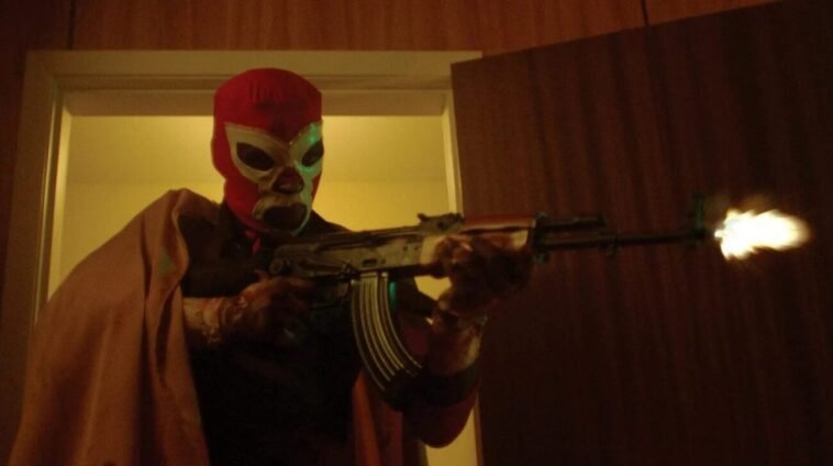 A luchador wrestler stands in a doorway, firing a machine gun