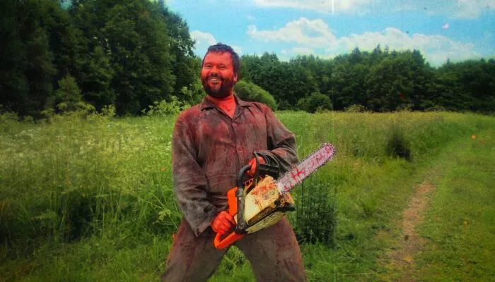 A man covered in blood stands in a green field, holding a chainsaw and smiling
