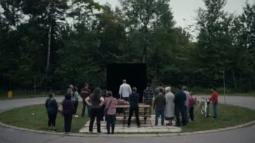 A gathering of people on a circular grassy area surrounded by roadway where a black square draws their gaze.