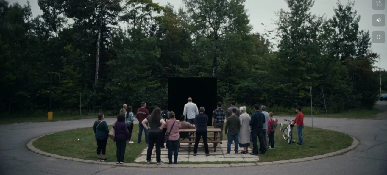 A gathering of people on a circular grassy area surrounded by roadway where a black square draws their gaze.