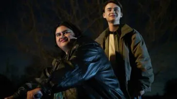 Two smiling young men on a bicycle, one steering, the other standing on pegs on the back wheel.