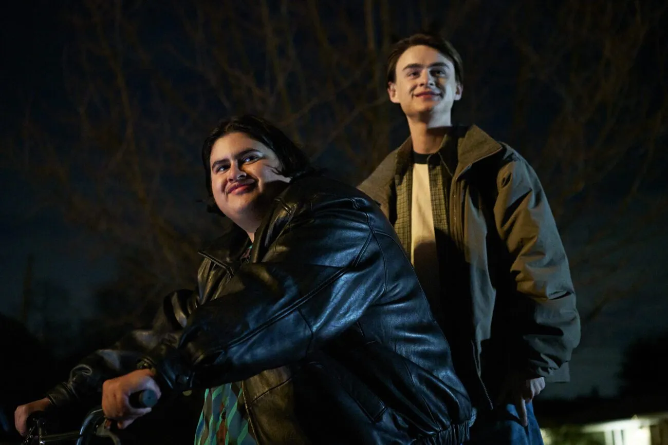 Two smiling young men on a bicycle, one steering, the other standing on pegs on the back wheel.