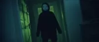 A man in a hoodie and a baby mask blocks a hallway in LISTEN CAREFULLY