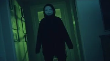 A man in a hoodie and a baby mask blocks a hallway in LISTEN CAREFULLY