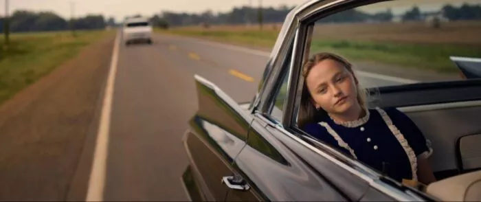 A young woman with her head leaned againse the window of a classic car, a white van follows behind.