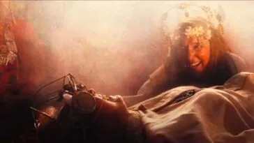 A woman in a flower headdress looms over a robed woman tied to a table.