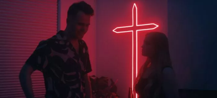 A man laughs while talking with a woman in the the glow of a neon cross.