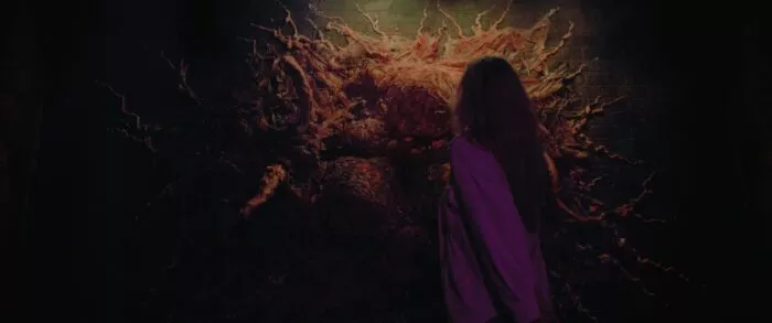 A woman stands in front of a visceral tentecle monster attached to a wall