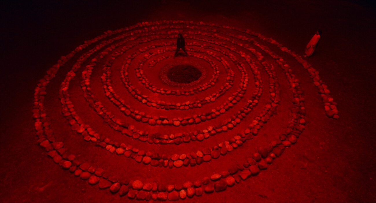 A stone spiral leads to a person in the center standing over a large hole in the ground in BOKSHI