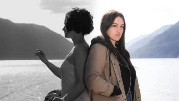 two women are seen back to back with a vast lake and mountains in the background, one is presented like a black and white photograph, the other in bright vivid color. THE LADY OF THE LAKE