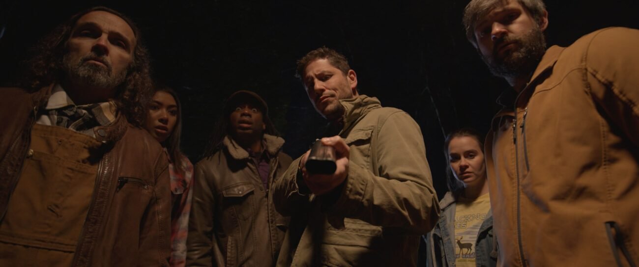 A man surrounded by a group of people holds a shotgun into the lens of the camera