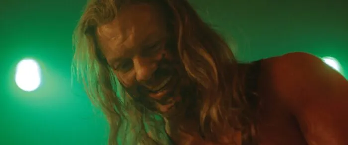 a man with long hair and facial scarring is seen smiling under a green light
