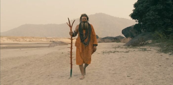 A shaman in a colorful robe and powder white face uses a walking stick to traverse a sandy beach in A Cursed Man