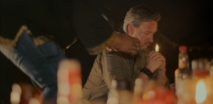 An unseen man's hands smear blood across another man's forehead as he sits among a large display of lit candles.