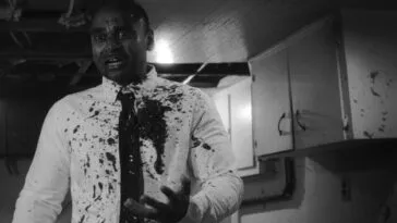 A black and white image of a man in a tie and white button down shirt standing in a half-finished basement covered in blood in HEAD LIKE A HOLE