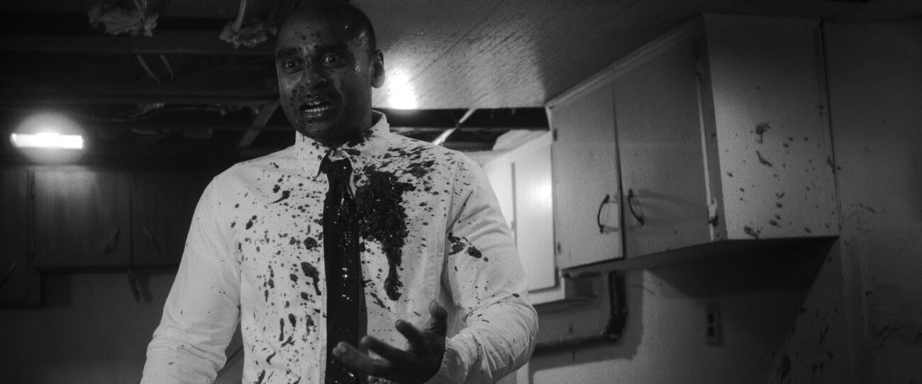 A black and white image of a man in a tie and white button down shirt standing in a half-finished basement covered in blood in HEAD LIKE A HOLE