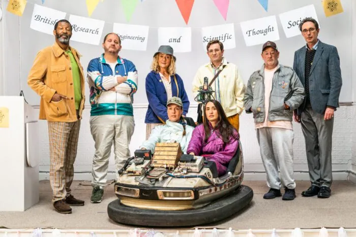 A group of men and women and stand around a bumper car where two women are seated.