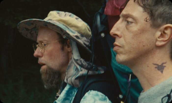 two men walking together, the one further away has a big camping backpack and a sun hat on.