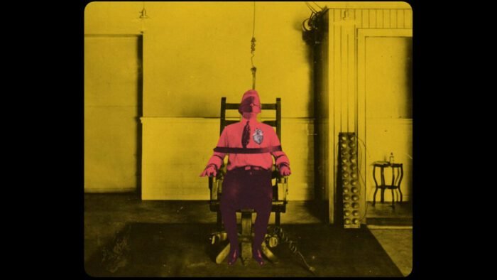 The image of a man tied to a chair who is highlighted in red against the yellow interior of the room and a white heart.