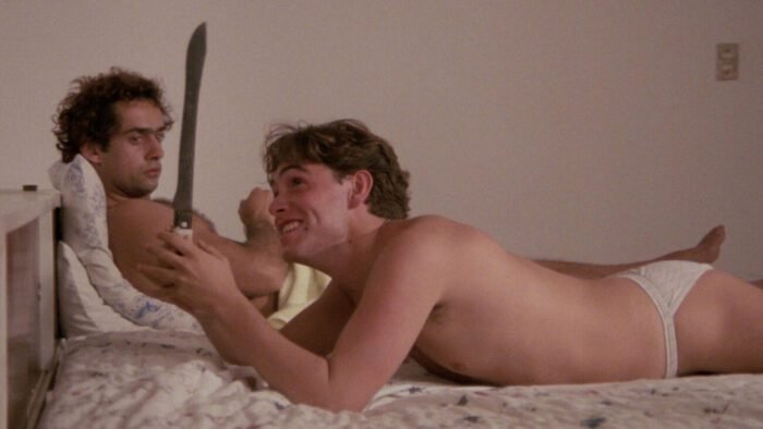 Two men laying on a bed in their underwear, the one closest to the camera is smiling while holding a large knife