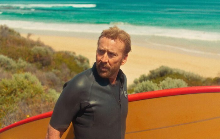 A man in a wetsuit on the beach carrying a surfboard toward the ocean looks over his shoulder in THE SURFER