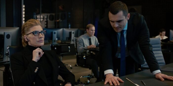 A woman sits in a chair at a desk listening to a man in the foreground speak while another man in the background looks at a computer screen.