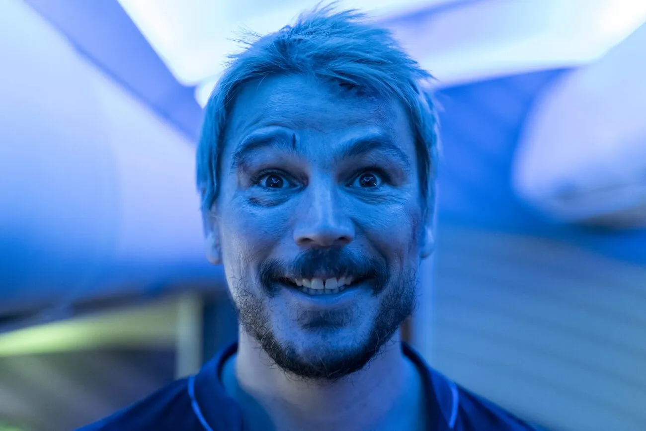A bleach blonde man with a black eye is seen smiling directly into the camera under an ambient blue light.