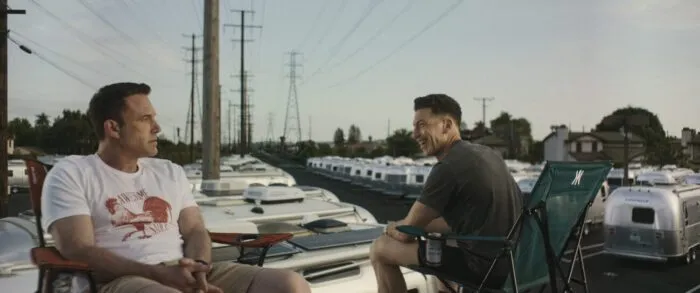 Two men sit together on the roof of a trailer, a sea of campers under power lines surrounding them