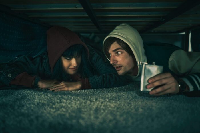Two people in hoodies lying on their bellies in a crawl space, one has a flask.