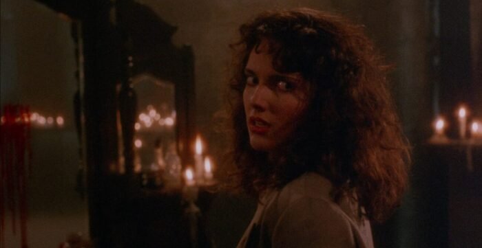 a woman turns away from a mirror adorned with lit candles looking over her shoulder into the camera