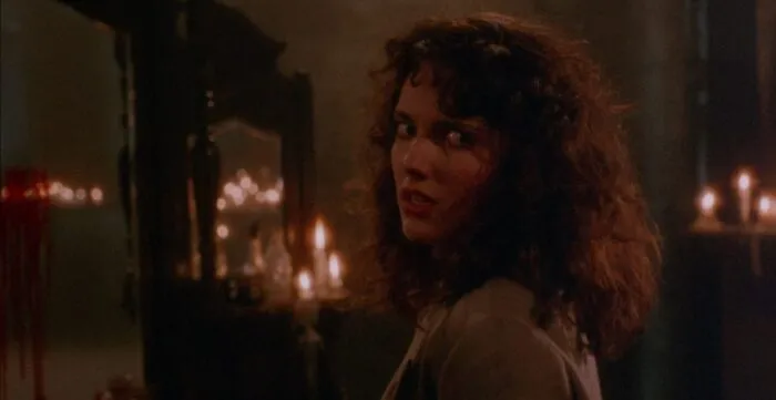a woman turns away from a mirror adorned with lit candles looking over her shoulder into the camera
