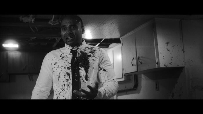 A black and white image of a man in a tie and white button down shirt standing in a half-finished basement covered in blood in HEAD LIKE A HOLE