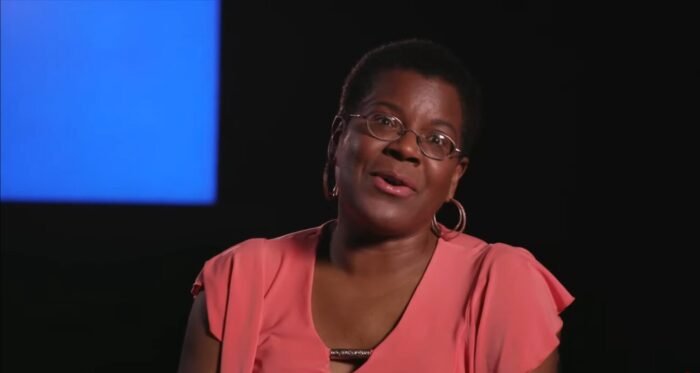 A woman in a pink shirt is speaking to the camera.