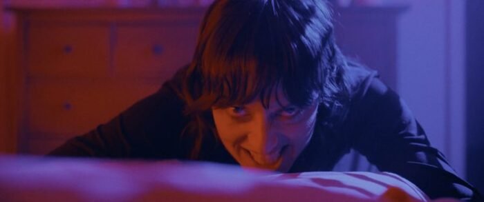 a woman looks diabolically into the camera from the end of a bed.