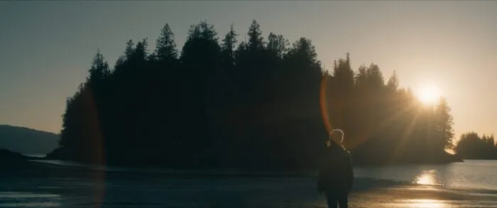 The Sun sits among the tree line of an island as a woman in a puffy coat and winter hat walks through the water at low tide in THE ISLAND BETWEEN TIDES