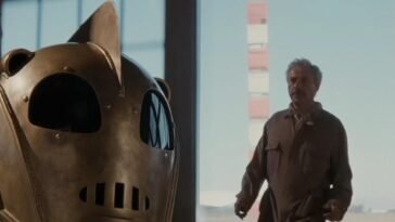 A older man walks into an airport hanger and looks at a bronze helmet sitting on a table