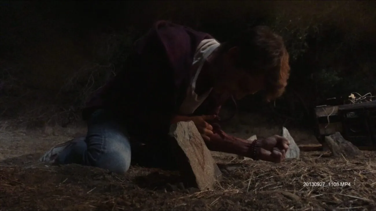 A man screaming in agony is seen crouched and inserting something into his arm on the edge of a circular rock formation.